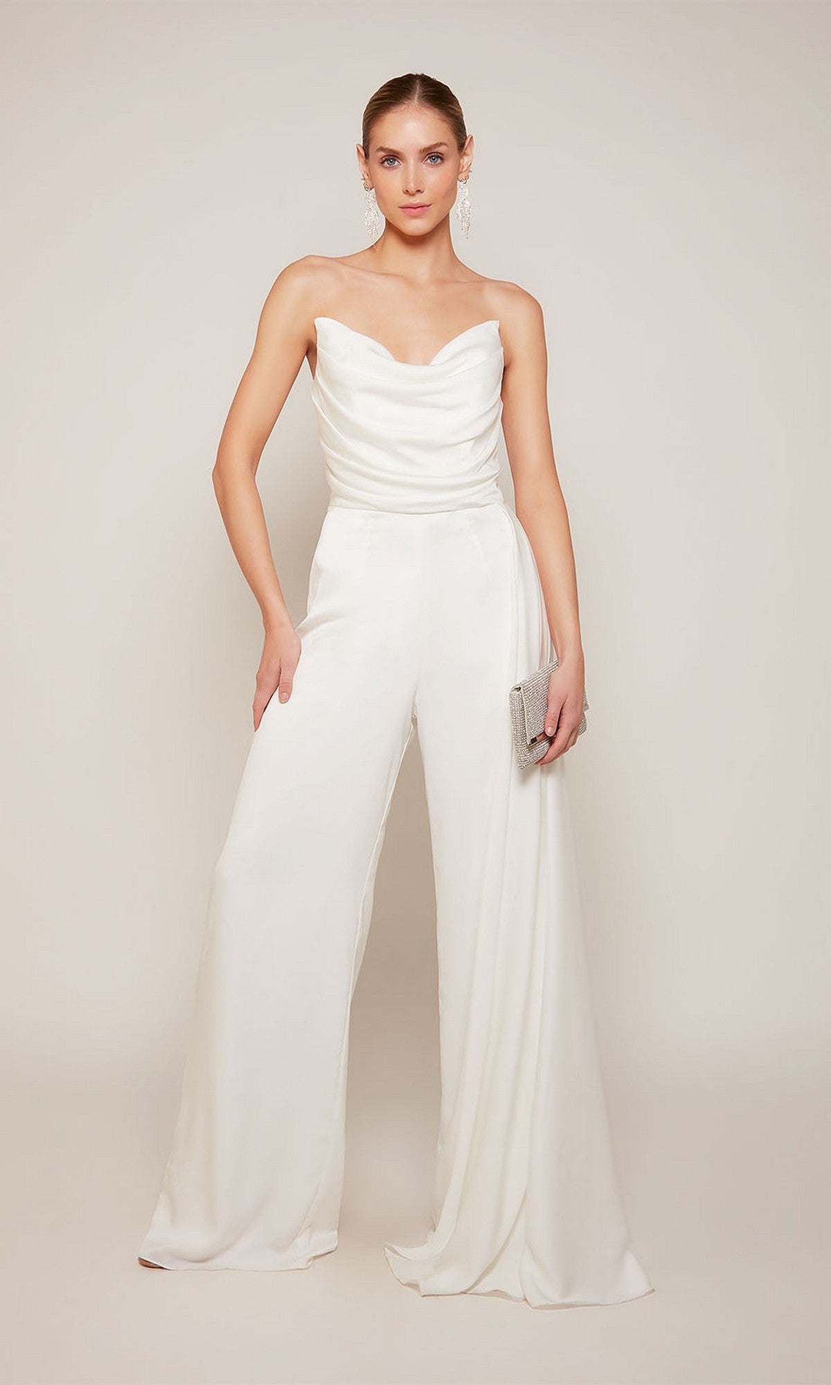 Woman wearing the Long Formal Jumpsuit: Alyce 70050, featuring a strapless neckline and wide-leg pants, holding a silver clutch against a plain light background.